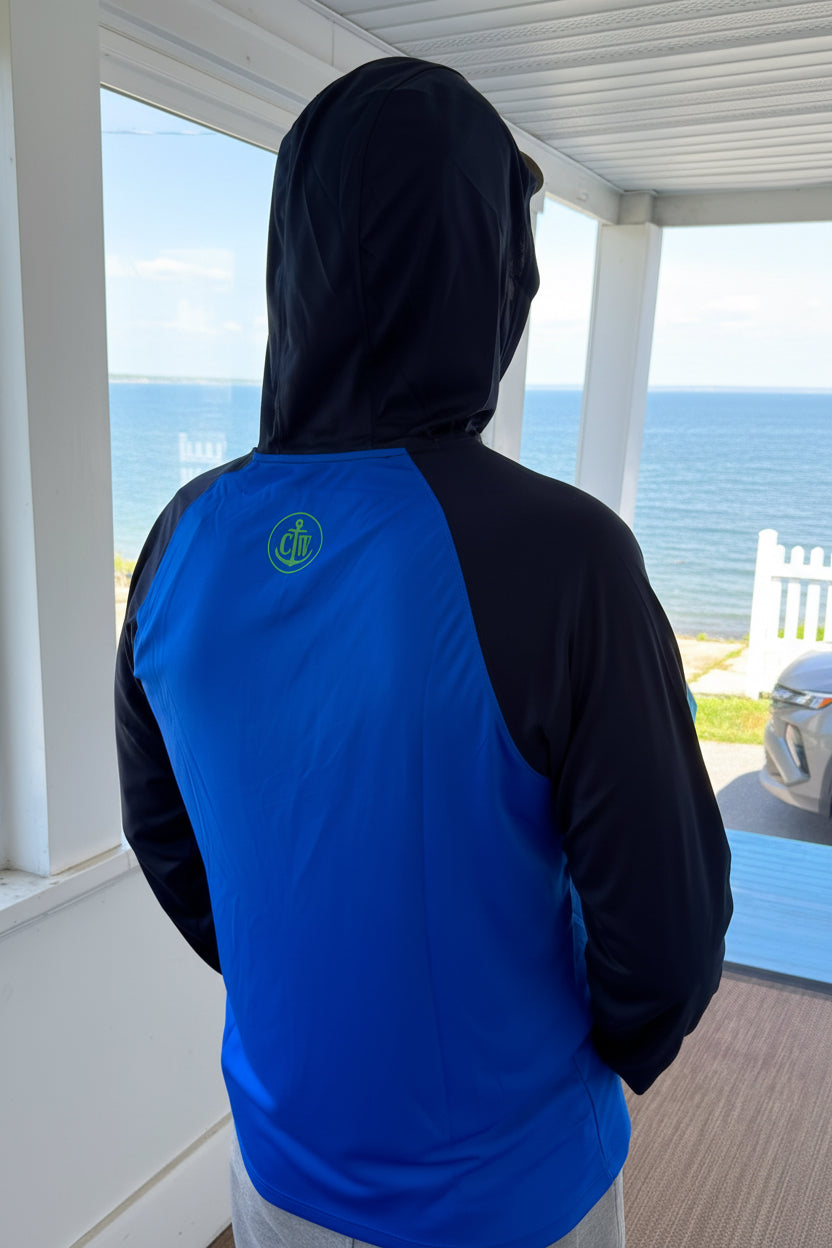 A person viewed from behind wearing a two-tone hooded athletic jacket with a bright blue back panel and black sleeves and hood, standing on a porch with the ocean visible outside.