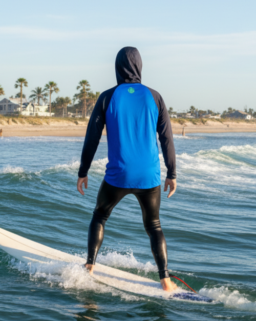A surfer viewed from behind wearing a bright blue and black hooded wetsuit while standing on a white surfboard in the ocean, with a beach and palm trees in the background under a clear blue sky.