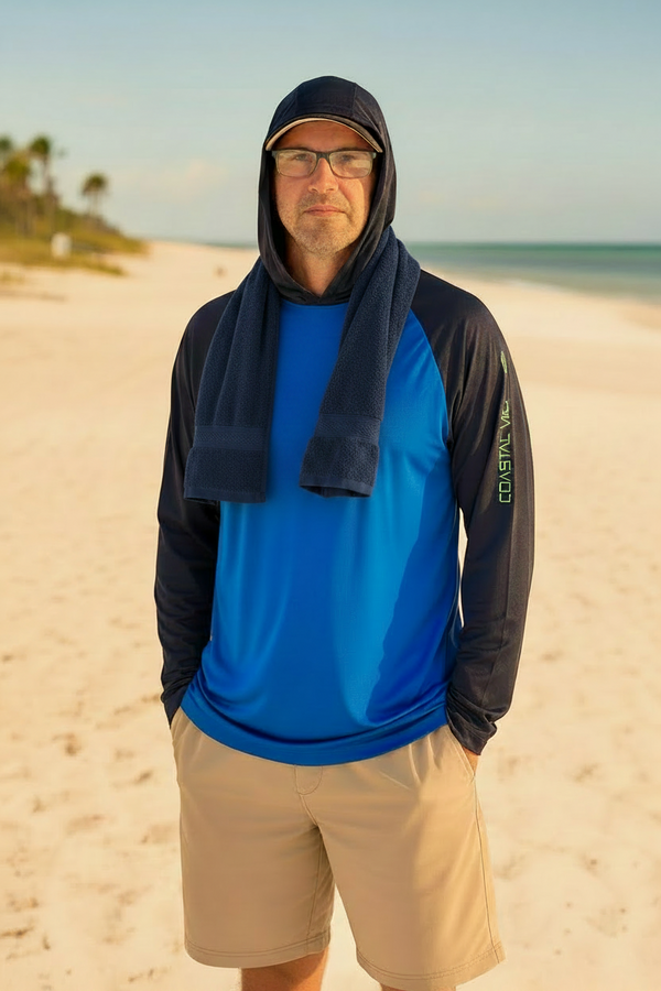 A man standing on a sandy beach wearing a blue and black long-sleeve performance shirt with a navy blue towel draped over his shoulders, khaki shorts, and a black cap.