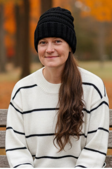 A woman wearing a black ribbed knit beanie and a cream sweater with black horizontal stripes, sitting on a wooden bench outdoors in a park with autumn trees in the background.