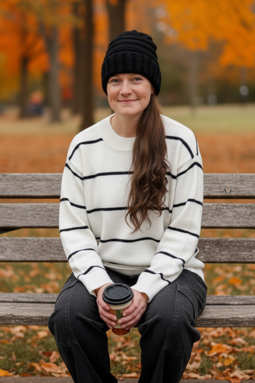 A woman sitting on a wooden park bench wearing a black knit beanie, a cream and black horizontally striped sweater, and dark gray pants, holding a takeaway coffee cup with autumn trees and fallen leaves in the background.