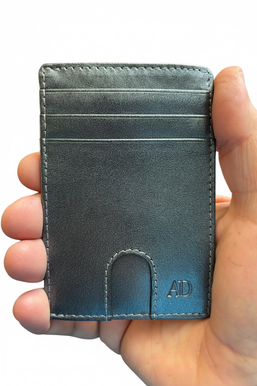 A hand holding a slim, minimalist cardholder wallet in a textured slate blue-gray leather. The cardholder features multiple card slots, precise stitching, and an embossed 'AD' monogram in the bottom right corner.