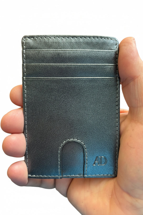 A hand holding a slim, minimalist cardholder wallet in a textured slate blue-gray leather. The cardholder features multiple card slots, precise stitching, and an embossed 'AD' monogram in the bottom right corner.