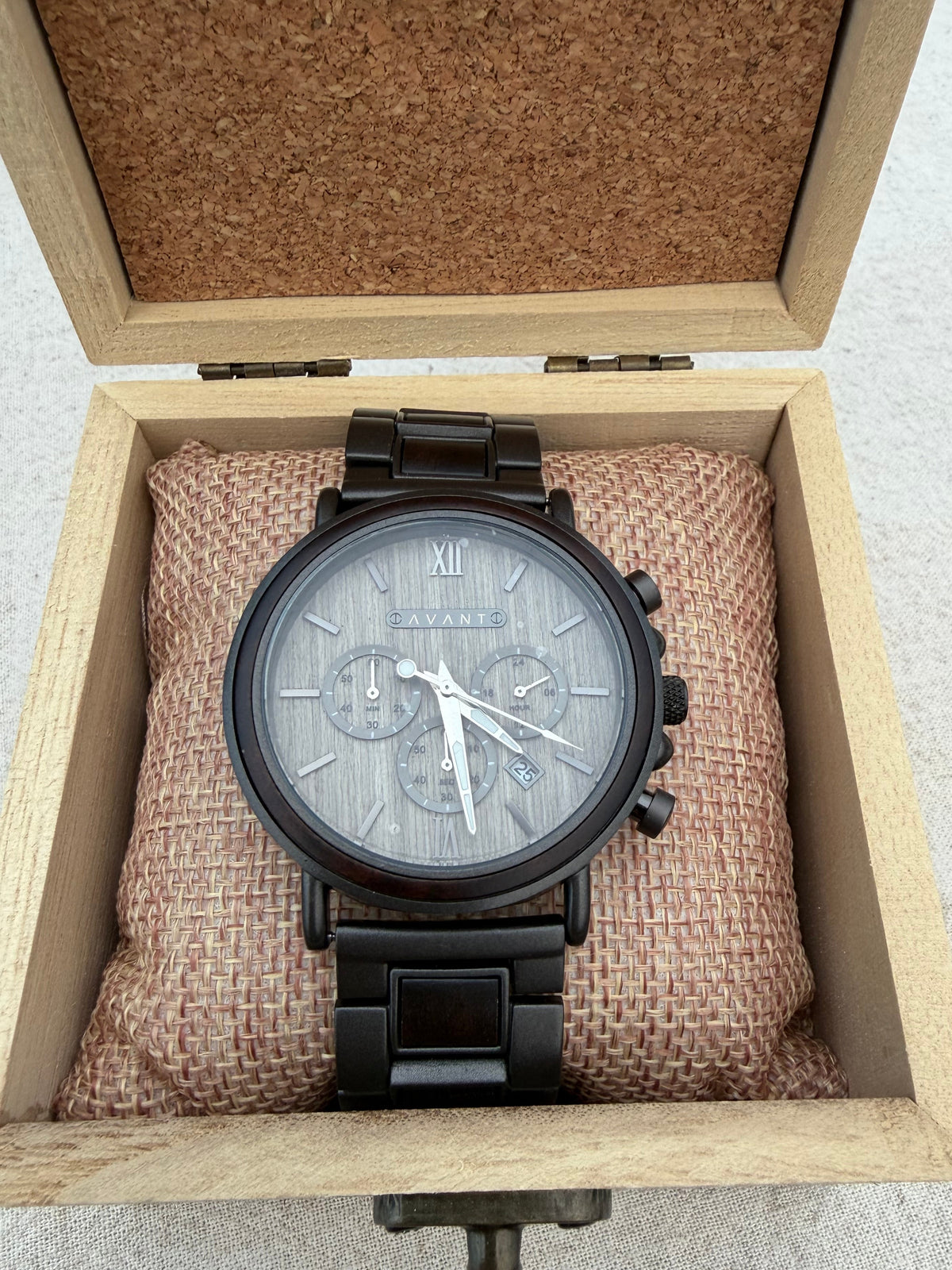 A black chronograph wristwatch with a gray textured dial and silver-tone hands, displayed inside an open light wood presentation box with a cork-lined lid and a brown fabric cushion.