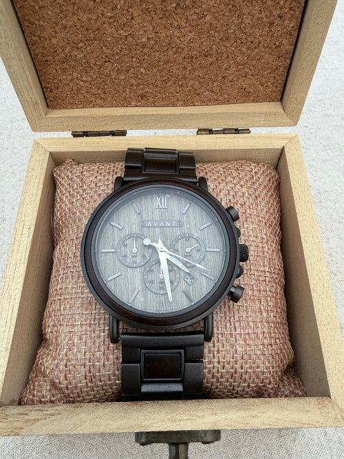 A black chronograph wristwatch with a gray textured dial and silver-tone hands, displayed inside an open light wood presentation box with a cork-lined lid and a brown fabric cushion.