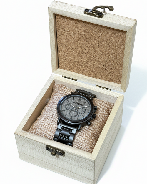 A black chronograph wristwatch with a black metal bracelet and gray dial, presented inside an open light wood gift box with a cork-lined lid on a white background.