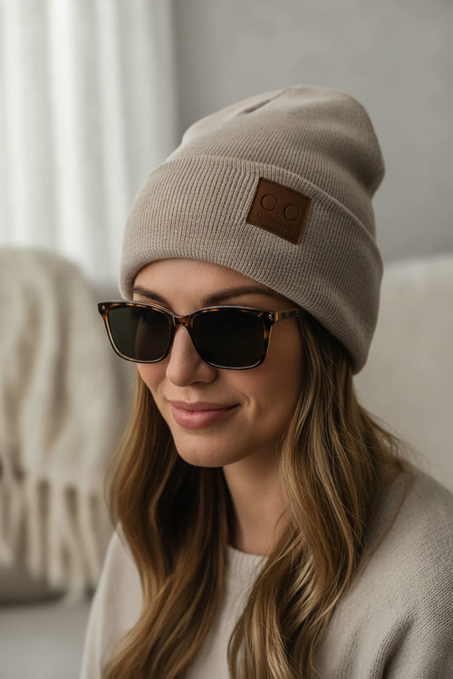 Woman wearing a beige beanie with a patch and dark sunglasses indoors.