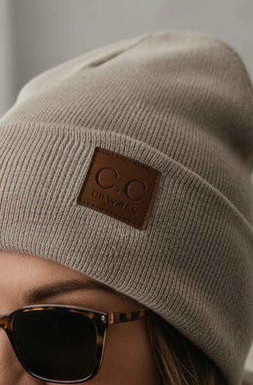 Close-up of a beige ribbed knit beanie with a brown leather patch reading 'C.C CLEWMAS' worn by a person wearing tortoiseshell sunglasses.