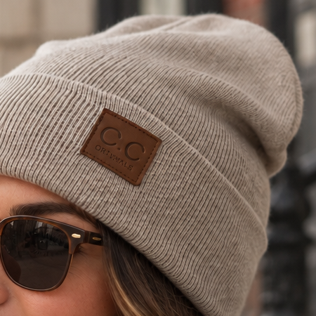 A woman wearing a beige knit beanie with a brown leather patch that reads 'CO. CLOTHING', posed outdoors against a blurred urban background.