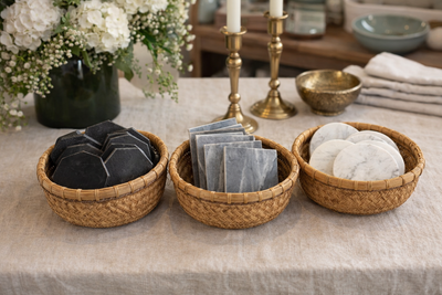 Three woven wicker baskets arranged on a linen tablecloth, holding black soap bars, gray stone tiles, and white marble coasters, with a vase of white flowers and brass candlesticks in the background.