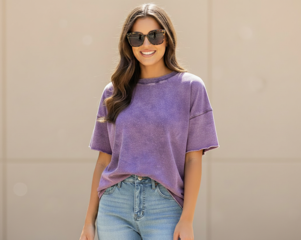 A woman wearing a purple short-sleeve crewneck t-shirt and light blue jeans, standing against a beige wall.
