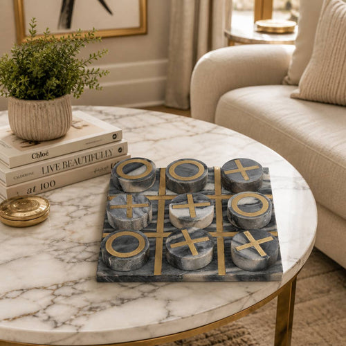 A marble-and-stone tic-tac-toe game board with round gray and white pieces sits on a round white marble coffee table in a styled living room setting.