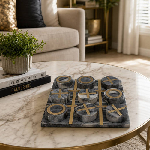 A round marble-top coffee table in a living room setting, featuring a dark gray and black stone tic-tac-toe game board with gold-colored cross and O shapes. The table is styled with a small potted green plant and a stack of books, with a beige sofa and window in the background.