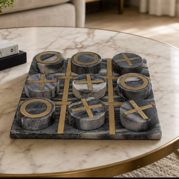 Family Game Sets A round marble-top coffee table in a living room setting, featuring a dark gray and black stone tic-tac-toe game board with gold-colored cross and O shapes. The table is styled with a small potted green plant and a stack of books, with a beige sofa and window in the background.