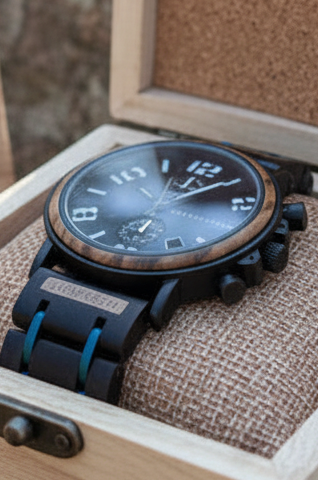 Wood Watch Pilot A black chronograph watch with a blue-accented strap rests inside an open light wood presentation box with a cork-lined lid, placed outdoors on a stone surface.