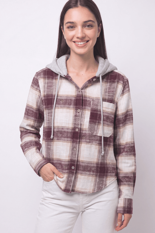 A woman modeling a long-sleeve hooded button-up shirt with a brown and beige plaid pattern and a light gray hood, paired with white pants against a light gray studio background.