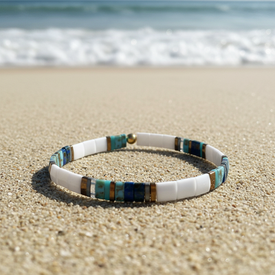 A beaded stretch bracelet resting on sandy beach sand with the ocean waves blurred in the background. The bracelet features white cylindrical beads, turquoise blue and teal stone beads, and small metallic gold spacer beads.