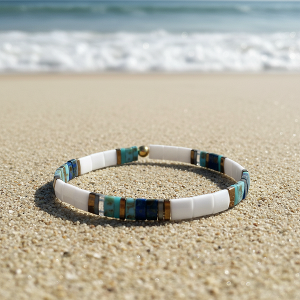 A beaded stretch bracelet resting on sandy beach sand with the ocean waves blurred in the background. The bracelet features white cylindrical beads, turquoise blue and teal stone beads, and small metallic gold spacer beads.
