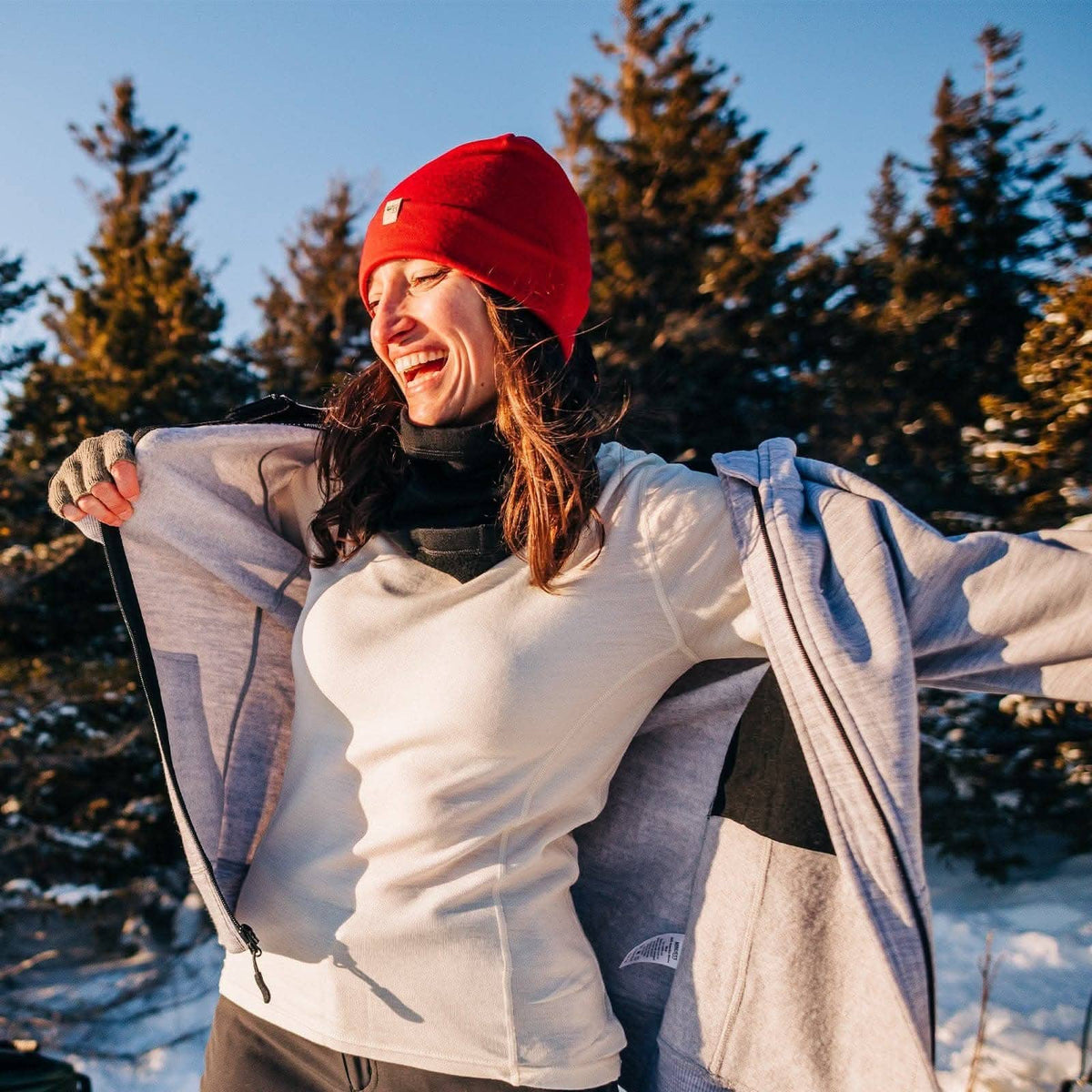 A woman outdoors in a snowy forest wearing a bright red knit beanie, a white long-sleeve top, and a light gray zip-up jacket, smiling with her arms outstretched.
