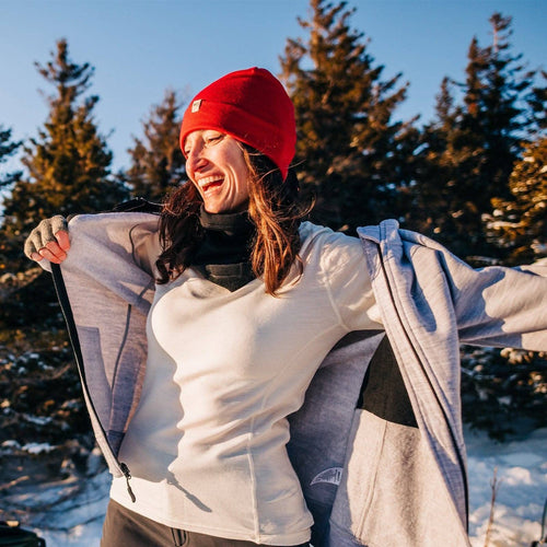 A woman outdoors in a snowy forest wearing a bright red knit beanie, a white long-sleeve top, and a light gray zip-up jacket, smiling with her arms outstretched.