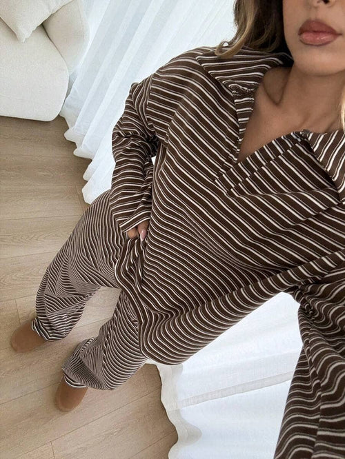 A woman wearing a brown and white striped pajama set with a collared long-sleeve top and matching pants, standing in a bright bedroom with light wood floors and white curtains.