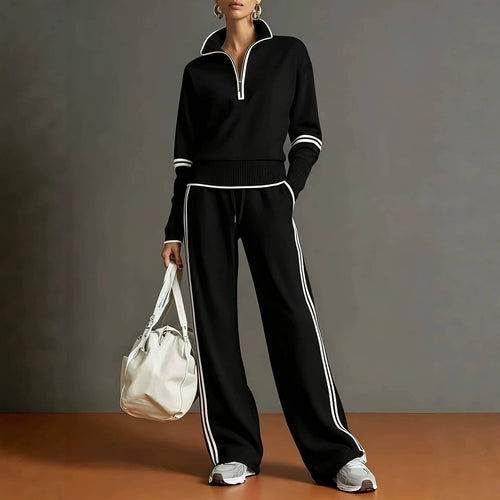 A model wearing a matching black half-zip sweatshirt and wide-leg track pants with white contrast stripes, holding a white bucket bag against a dark gray studio background.