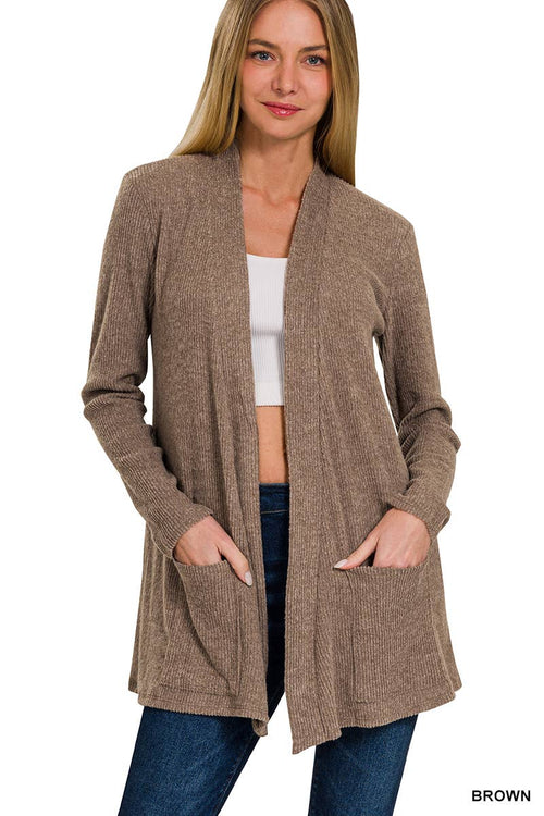 A woman modeling a brown open-front cardigan with long sleeves and front pockets, worn over a white crop top and blue jeans, against a plain white background.