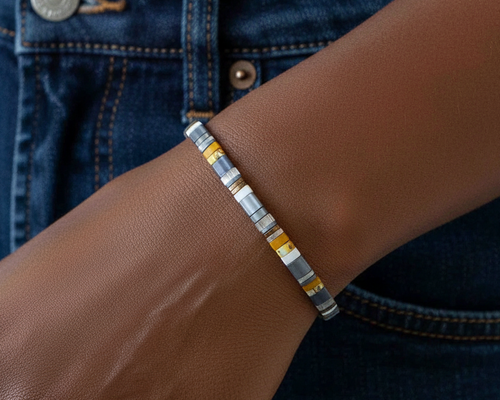 A close-up of a person's wrist wearing a beaded stretch bracelet with alternating translucent gray, white, and yellow beads, shown against dark blue denim jeans.