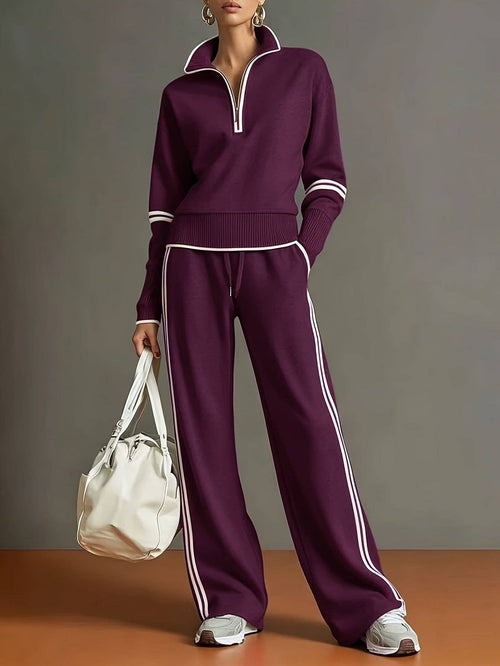 A model wears a matching deep purple tracksuit with white stripe accents, featuring a half-zip pullover and wide-leg pants, holding a white tote bag against a gray studio background.