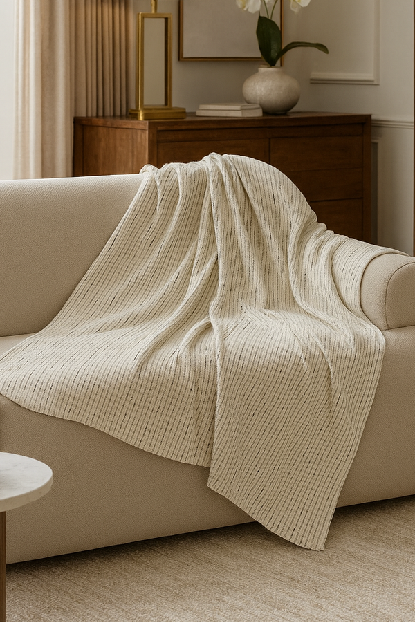 A cream-colored ribbed knit throw blanket is draped over a beige sofa in a warm, modern living room setting.