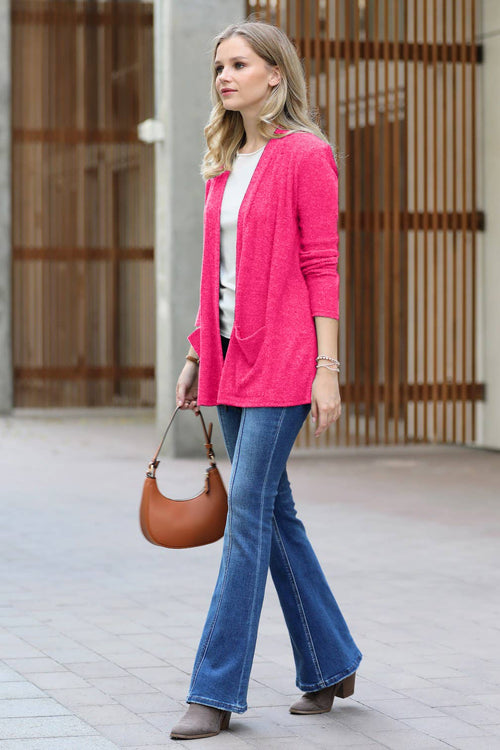 A woman walking outdoors wearing a bright pink open-front cardigan over a white top, blue flared jeans, and brown ankle boots, while carrying a brown leather handbag.