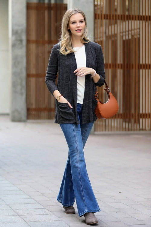 A woman wearing a dark gray open-front cardigan over a white top, blue flared jeans, and brown ankle boots, carrying a brown leather shoulder bag in an outdoor urban setting.