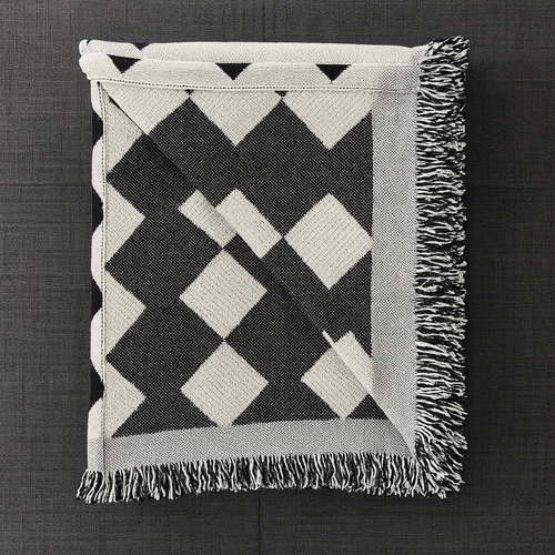 A folded woven throw blanket with a black and off-white geometric diamond pattern and fringed edges, laid flat on a dark textured background.