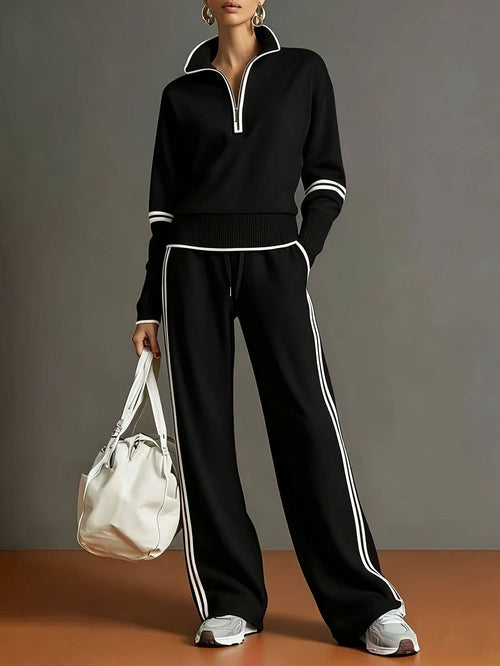 A model wearing a matching black knit tracksuit with white contrast trim, featuring a half-zip long-sleeve top and wide-leg pants with white side stripes. She is holding a white bucket bag and wearing white sneakers, set against a dark gray studio background with a warm brown floor.