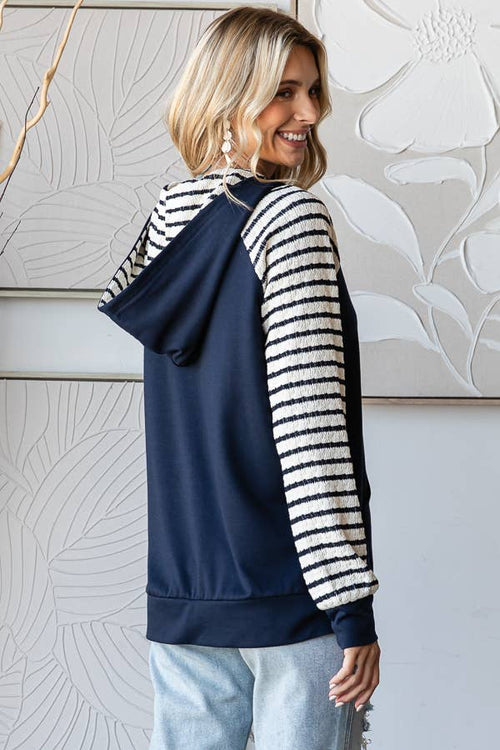 A woman wearing a navy blue hoodie with textured cream and navy striped raglan sleeves, shown from a three-quarter back view against a decorative light gray wall.