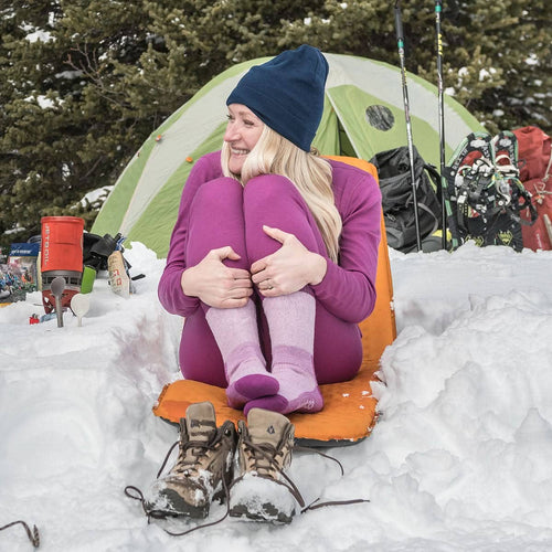 Person sitting on an orange sleeping pad in the snow with a green tent and camping gear in the background.