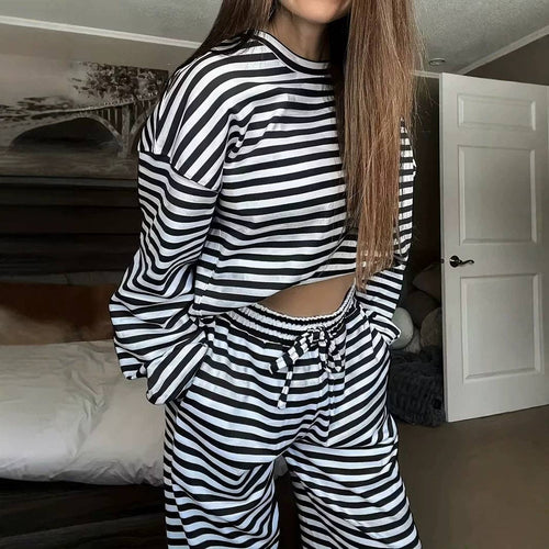 A woman wearing a matching black and white horizontally striped lounge set, consisting of a cropped long-sleeve top and drawstring sweatpants, standing in a bedroom.