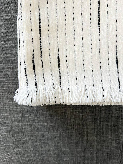 A close-up of a white woven textile with black vertical dashed stripes and fringed edges, laid on a dark gray textured surface.
