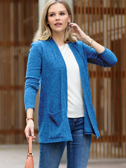 A woman wearing a heathered blue open-front cardigan over a white top and blue jeans, holding a peach handbag, set against a wooden slatted background.