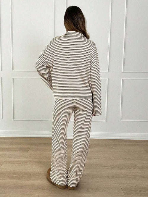 A woman viewed from behind wearing a matching beige and white striped pajama set with a long-sleeve top and wide-leg pants, standing in a room with white paneled walls and light wood flooring.