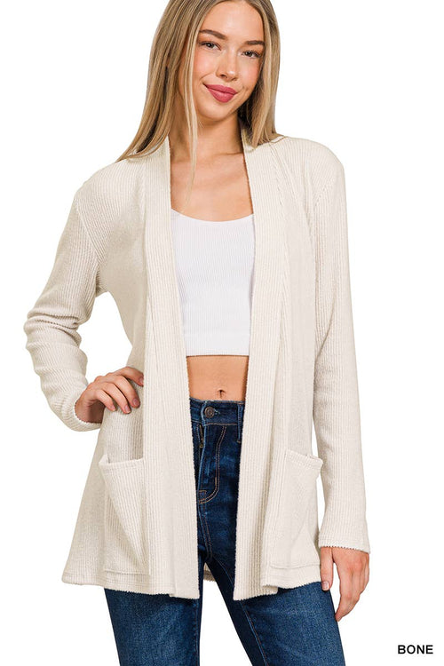 A woman modeling a ribbed knit open-front cardigan in a warm off-white or cream shade, labeled 'BONE'. She is wearing a white crop top and blue jeans against a plain white background.