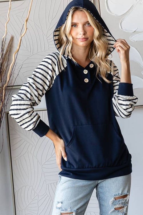 A woman wearing a navy blue raglan hoodie with black and white striped sleeves and hood trim, paired with light blue ripped jeans, standing against a light gray patterned wall.