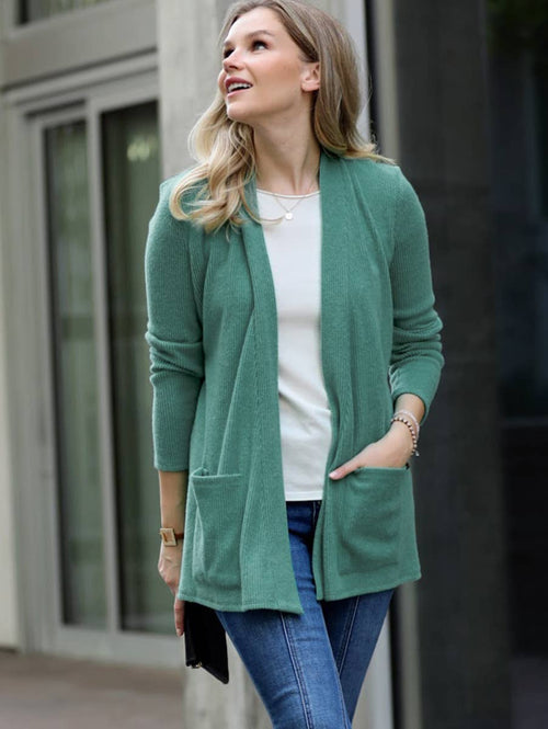 A woman wearing a sage green open-front cardigan over a white top and blue jeans, standing outdoors in front of a modern building with glass doors.