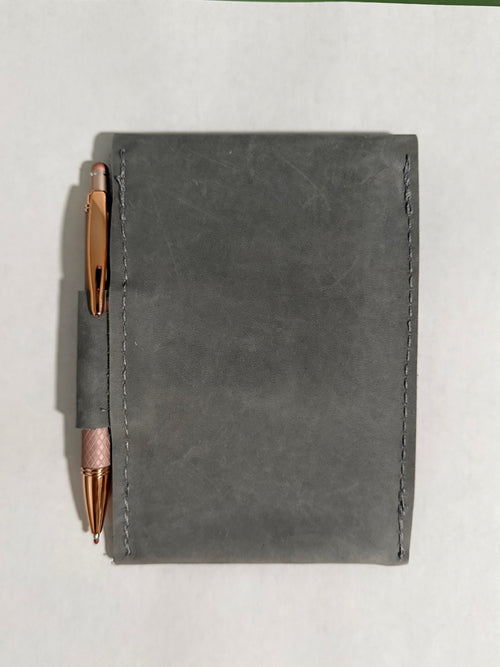 A dark gray leather notebook cover with a pen loop holding a rose gold pen, shown from a top-down view on a light gray background.
