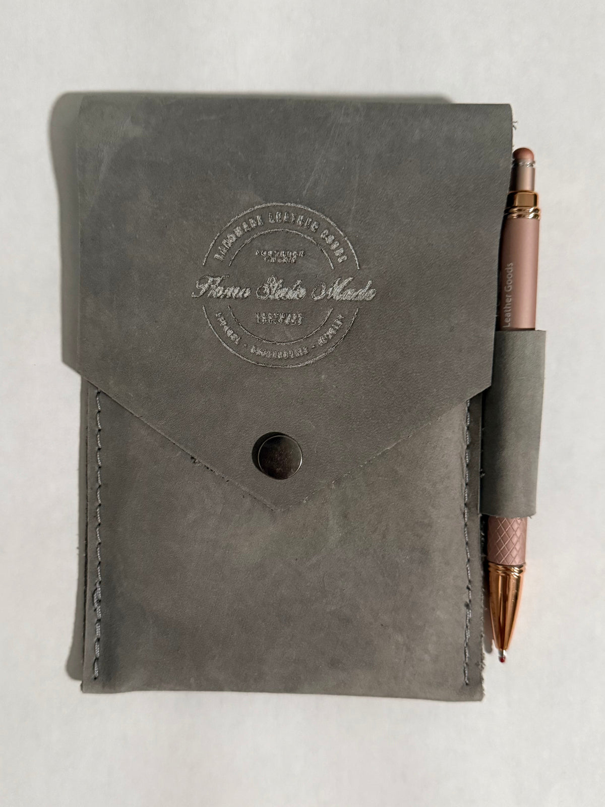 A dark gray leather notebook cover with a flap closure and a debossed circular logo, shown with a rose gold pen tucked into a side loop on a white background.
