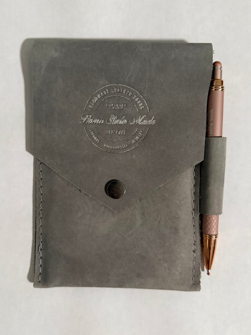 A dark gray leather notebook cover with a flap closure and a debossed circular logo, shown with a rose gold pen tucked into a side loop on a white background.