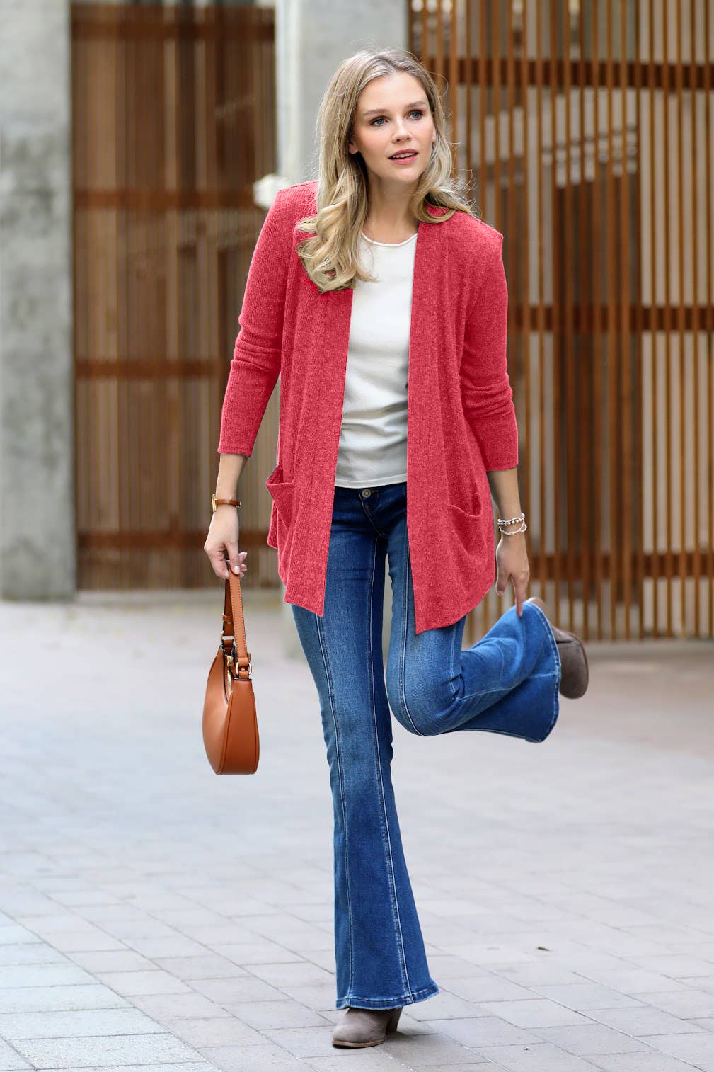A woman wearing a coral pink open-front cardigan, blue flare jeans, and a white top walks outdoors while holding a brown leather handbag.