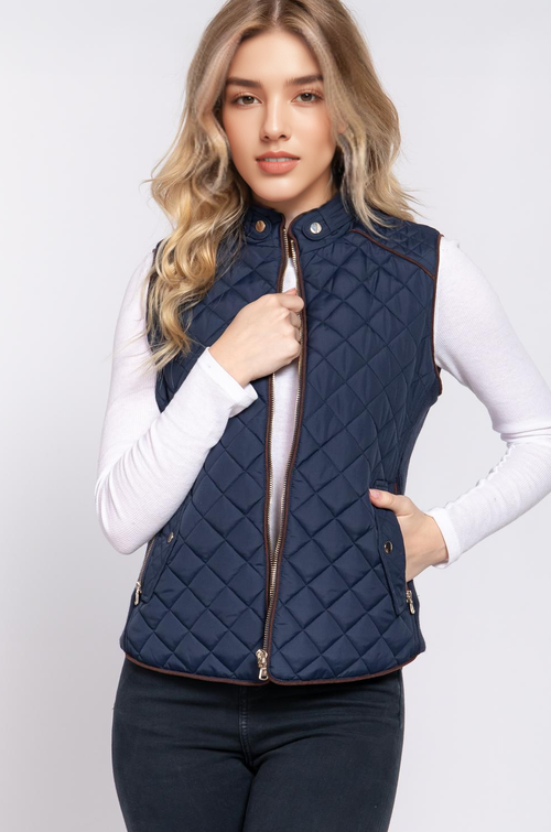 A woman modeling a navy blue diamond-quilted puffer vest with brown trim over a white long-sleeve shirt and dark jeans, set against a plain light gray studio background.