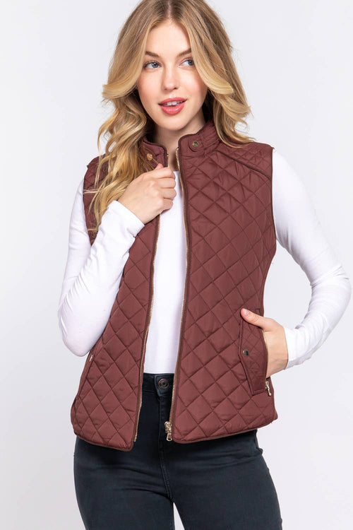 A woman wearing a quilted sleeveless vest in a dusty rose-brown color over a white long-sleeve top and black jeans, photographed against a light gray studio background.