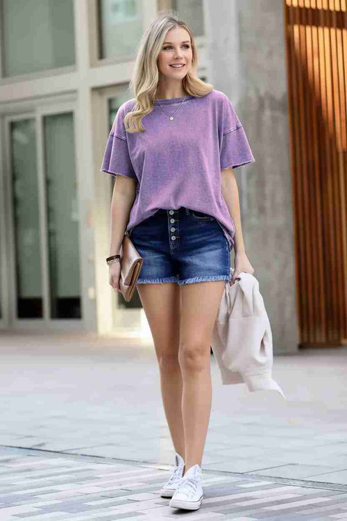 Woman wearing a purple oversized t-shirt and denim shorts standing on a street.
